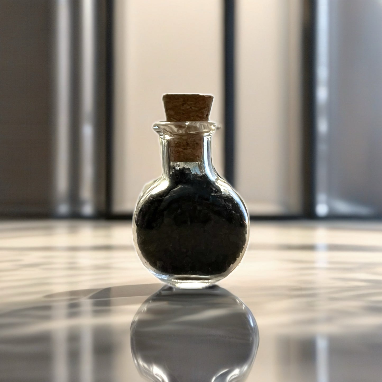 Small glass bottle with cork lid containing a black salt, on a reflective surface.